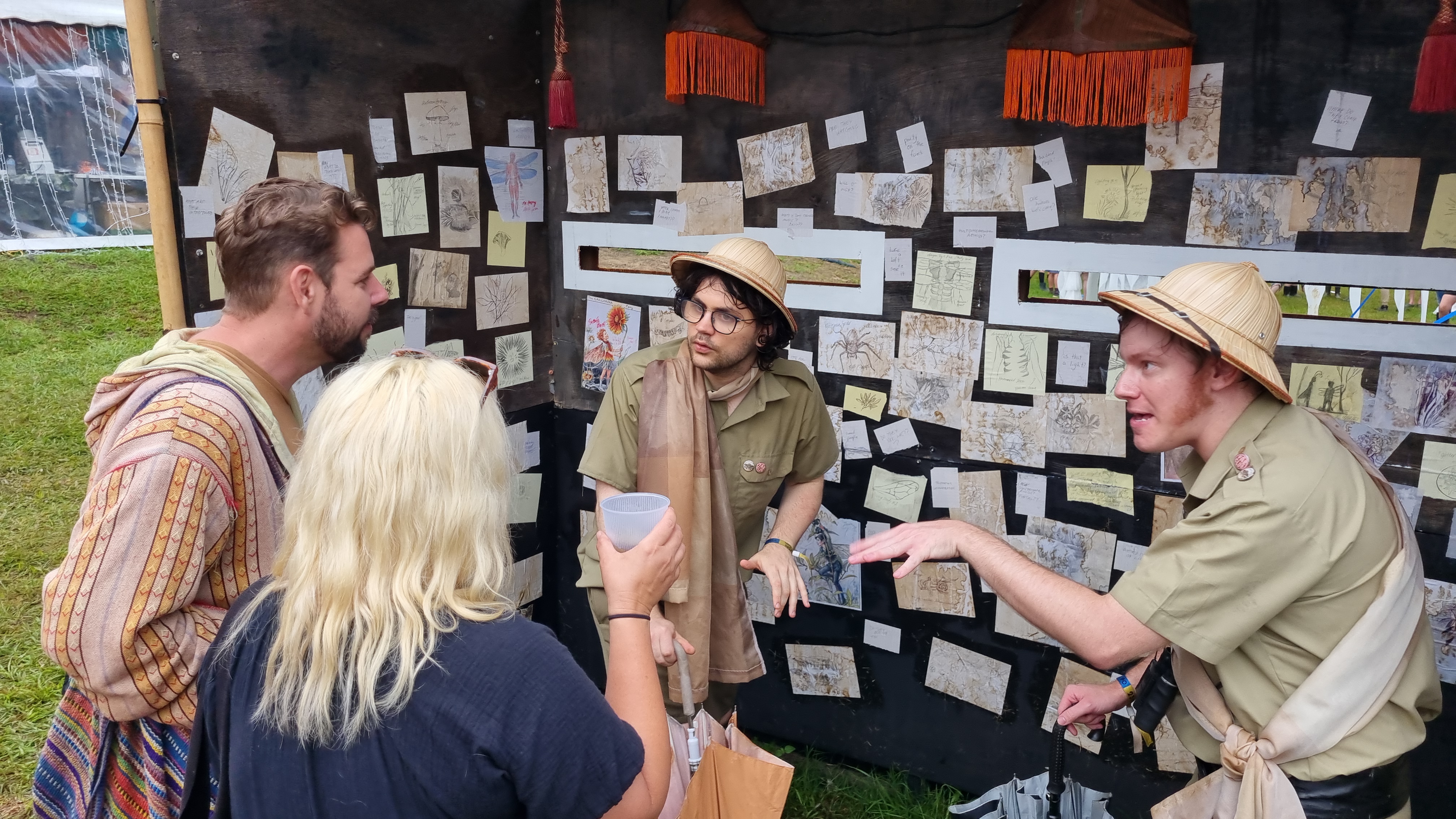 Two Faleontologists dressed in safari gear explain the Fae to two memebers of the public. They stand in front of a booth covered in drawing of fae creatures.